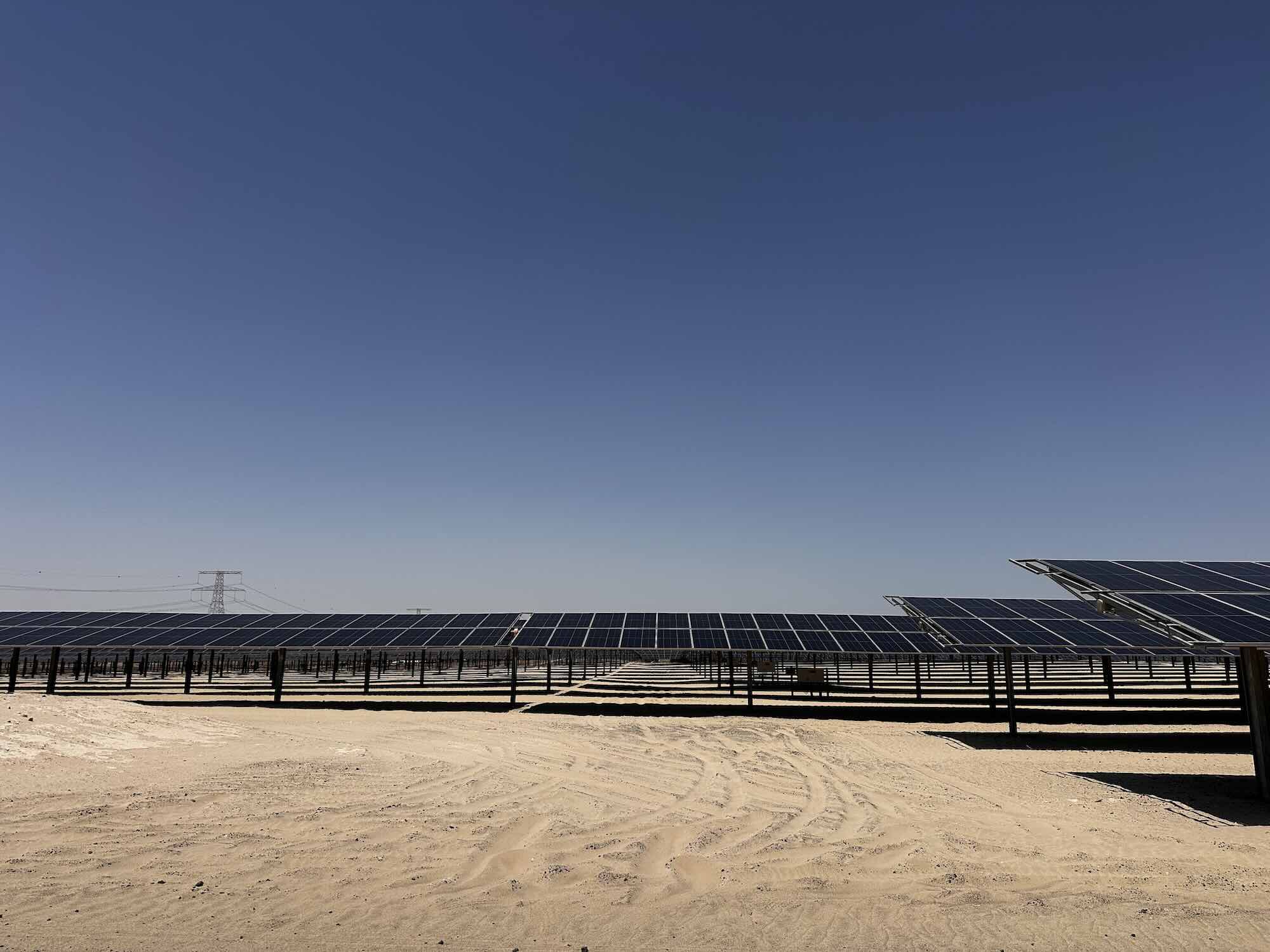 A large-scale solar power plant with rows of photovoltaic panels in a desert environment under a clear blue sky.